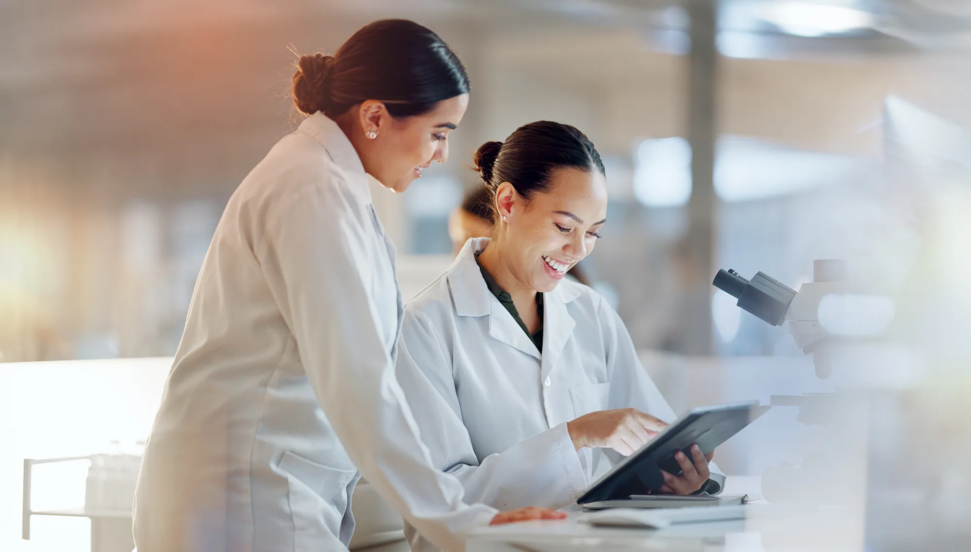 Two scientists in lab coats look at a tablet together next to a microscope.