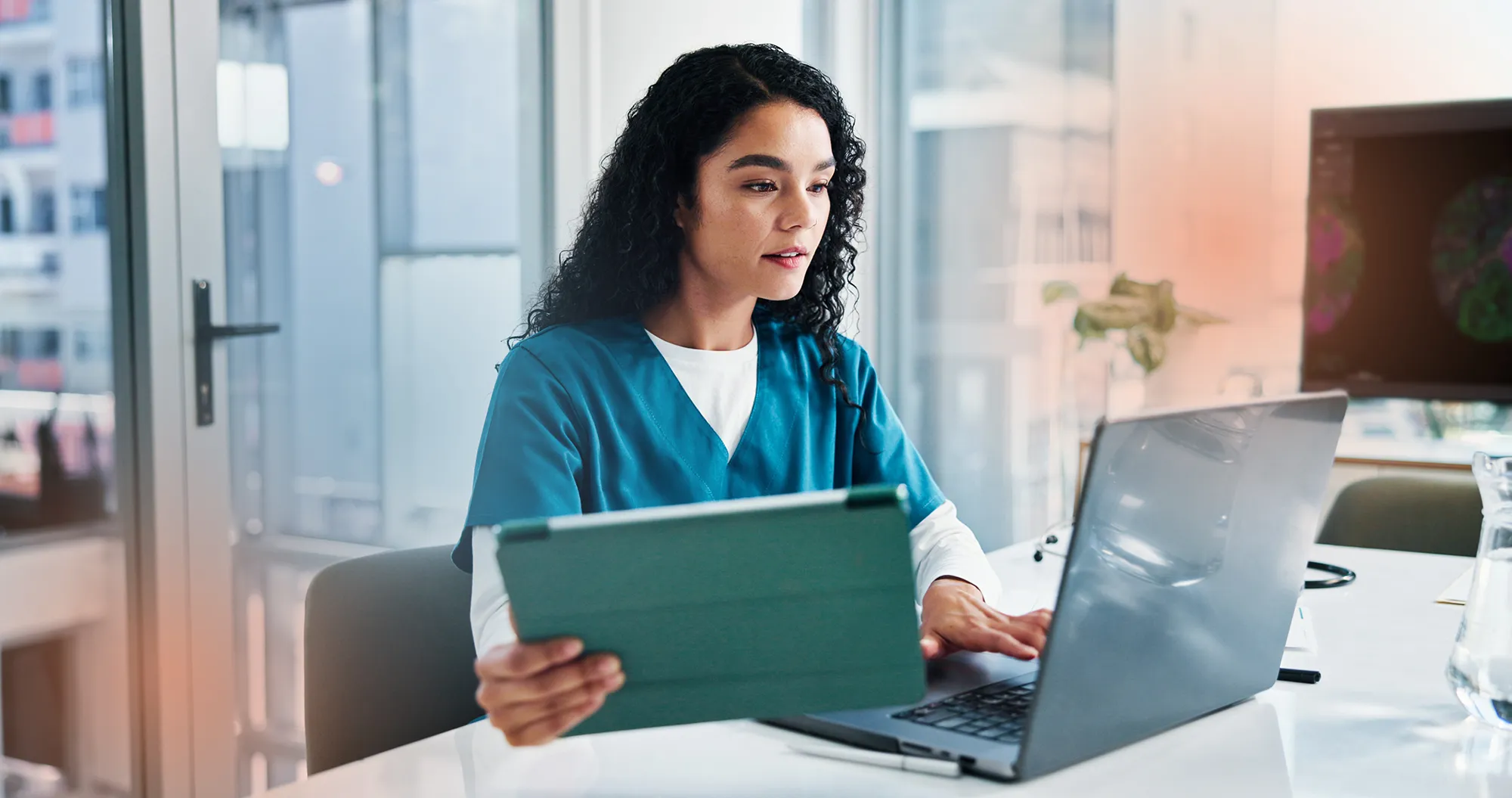 A scientist in scrubs holds a tablet next to a laptop at a desk.